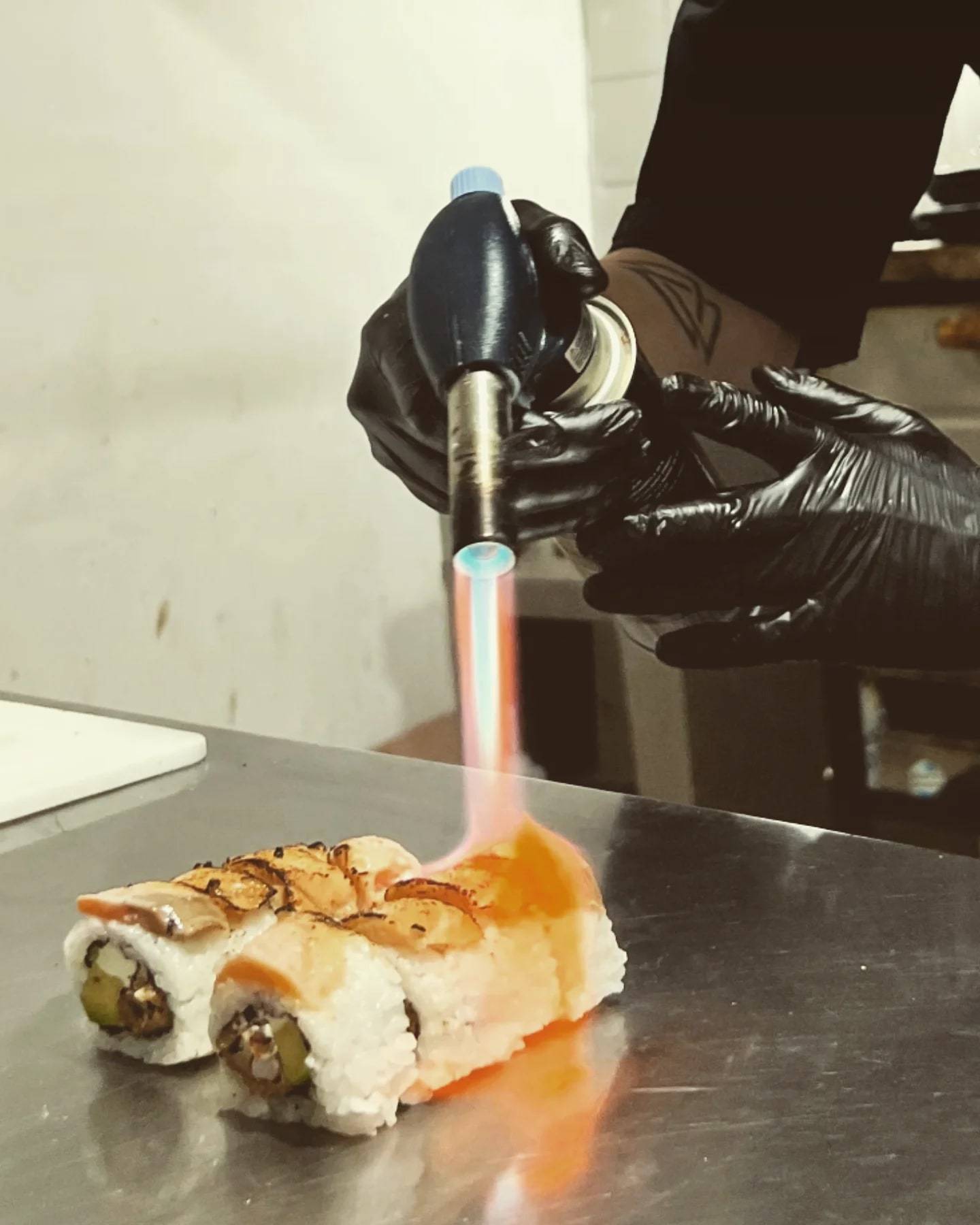 Manos con guantes negros sopletean un roll de sushi sobre una mesada de acero
