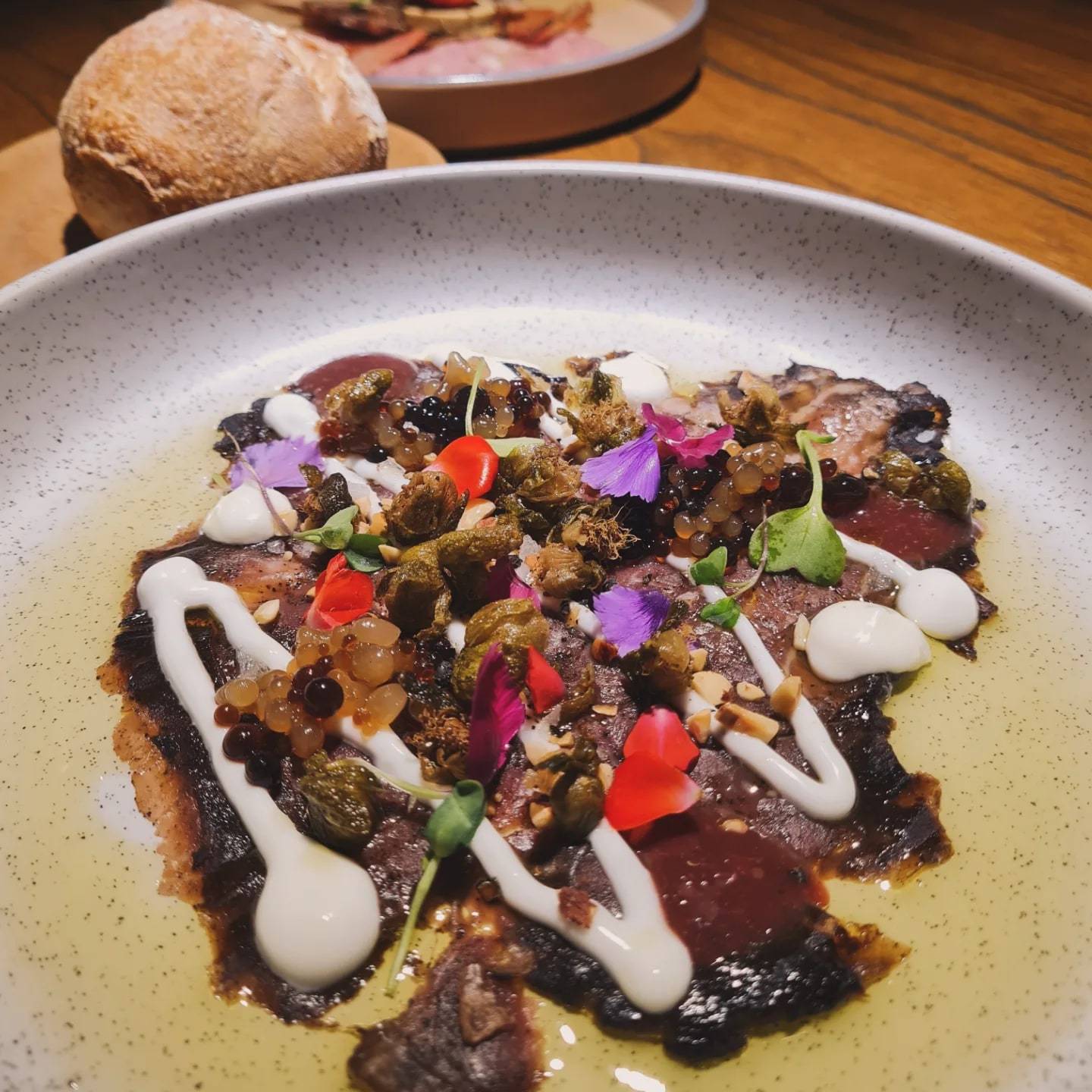 Láminas de carne cubiertas de alcaparras, perlas, brotes y flores violetas, con trazos de emulsión blanca en un plato moteado