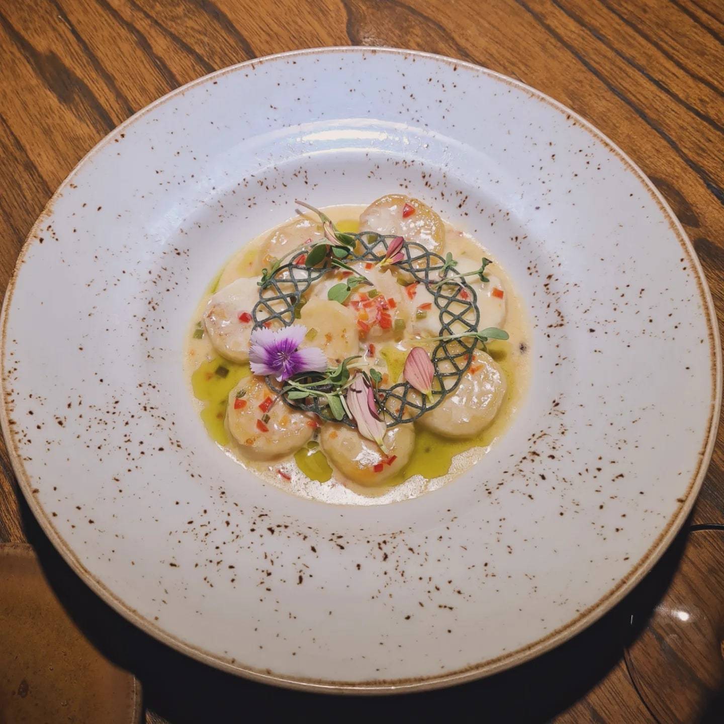 Láminas de mariscos en círculo sobre aceite verde, coronadas con una teja calada, brotes y flores, en un plato hondo moteado